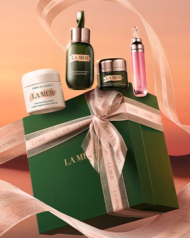 Green La Mer gift box with gold ribbon tied in a bow, with additional ribbon floating around and la mer products sitting on top.